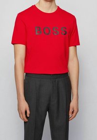 Person iført en rød T-shirt med "BOSS" i sorte bogstaver, parret med grå plisserede bukser, der står mod en ensfarvet lys baggrund.