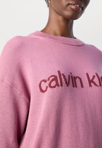 Gros plan d'une personne portant un pull en maille rose avec le texte "calvin klein" en rouge sur la poitrine, montrant uniquement les lèvres et le cou.