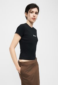 Νεαρό άτομο με κοντά σκούρα μαλλιά, φορέοντας μαύρο εφαρμοστό μπλουζάκι και καφέ φούστα, στέκεται με το ένα χέρι στην τσέπη μπροστά σε λευκό φόντο.