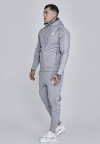 Svetlosivá mikina s kapucňou a zodpovedajúce jogger nohavice z hladkej tkaniny, s bočnými vreckami na zips a logom na hrudi a bočných švoch. Biele tenisky.