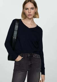 Mornarsko modra dolgosleeve V-izrez majica iz mehkega blaga, s sproščenim krojem, nosi se z temno sivo visoko pasom in črno torbo čez ramo.