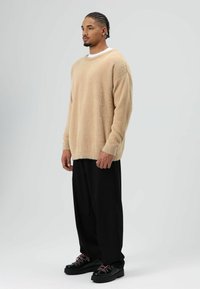 Beige gebreide trui met lange mouwen, gedragen over een wit T-shirt, gecombineerd met zwarte losse broek en zwarte sneakers met roze accenten.