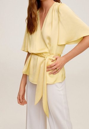 Żółta kopertowa bluzka z falbankowymi rękawami, wiązana w talii; wykonana z gładkiej tkaniny. Połączona z białymi spodniami, ukazującymi miękki drapowanie i przepływ materiału.