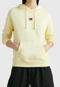 Sudadera amarilla hecha de tela suave, con un bolsillo frontal, capucha con cordón y un pequeño parche de logotipo en rojo y azul oscuro en el pecho.