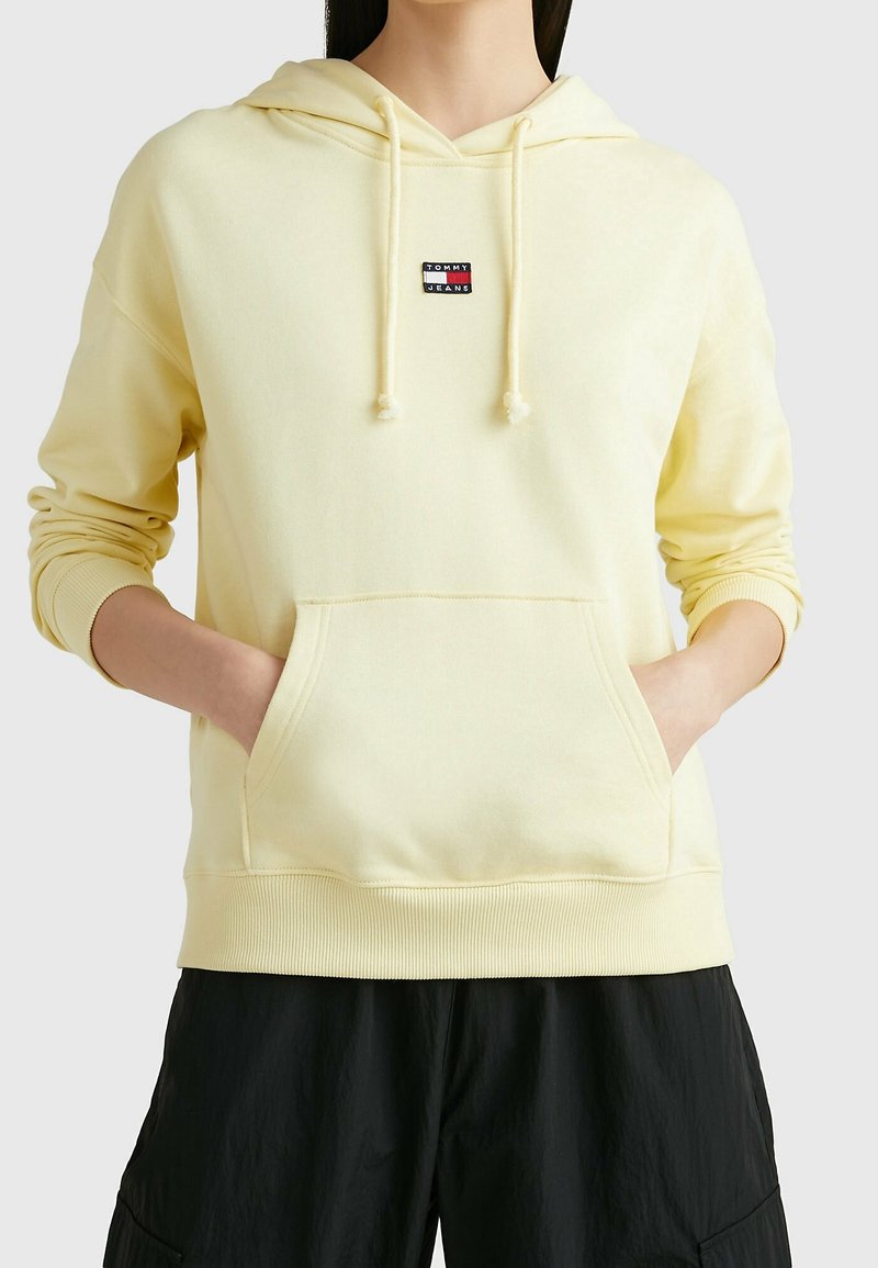 Sudadera amarilla hecha de tela suave, con un bolsillo frontal, capucha con cordón y un pequeño parche de logotipo en rojo y azul oscuro en el pecho.