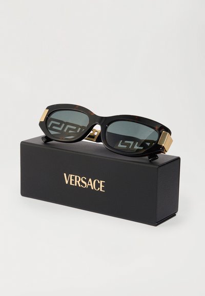 Versace solbriller med skildpaddefarvet stel, grønne linser, gyldne hardware-detaljer og et karakteristisk logodesign, placeret på en sort boks.