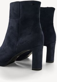 Navy modré semišové kotníkové boty s hladkým povrchem, zaoblenou špičkou a masivním vysokým podpatkem. Disponují minimálním šitím a elegantním designem.