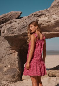 Abito rosa con motivo a fantasia e orlo a balze, caratterizzato da spalline annodate e una vestibilità comoda, su uno sfondo di spiaggia rocciosa.