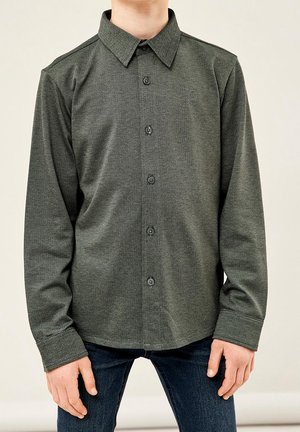 Junge trägt ein dunkelgrünes, langärmliges Hemd mit Knöpfen und dunkelblaue Jeans, steht mit entspannten Armen vor einem einfarbigen Hintergrund.