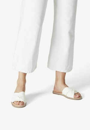 Pieds portant des sandales blanches plates à brides croisées, associées à un pantalon blanc large et raccourci, sur un fond blanc uni.
