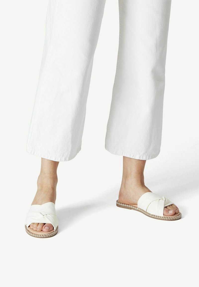 Pieds portant des sandales blanches plates à brides croisées, associées à un pantalon blanc large et raccourci, sur un fond blanc uni.