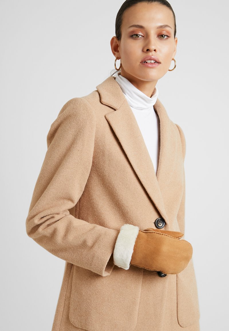 Beige wollen mix jas met notch revers, enkele knoopsluiting en twee voorkosten. Bruine suède wanten met witte faux bont voering.