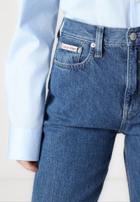 Modre kavbojke z visokim pasom, ki imajo rdeče-bel etiket Calvin Klein na žepu, z gladko teksturo in klasičnim pet-žepnim dizajnom.