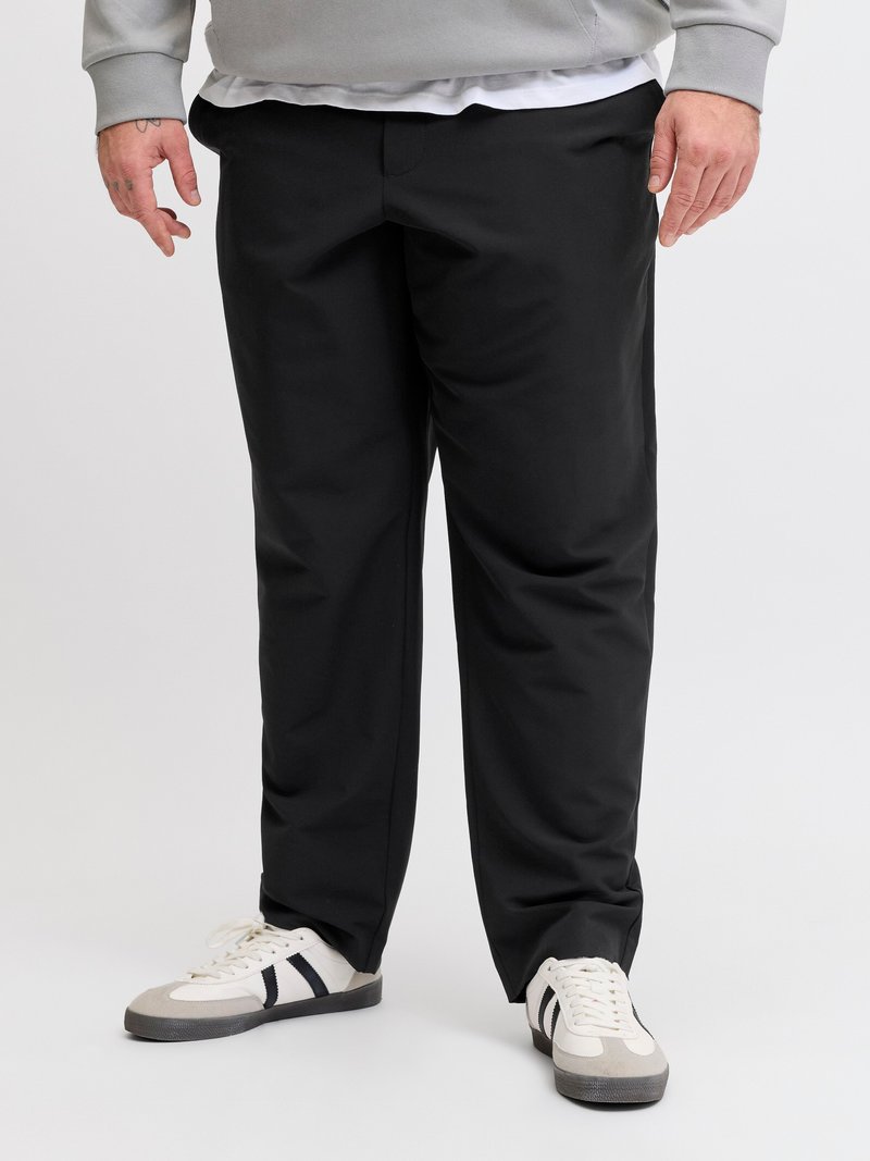 Uomo che indossa pantaloni neri a gamba dritta, sneakers bianche con strisce nere e felpa grigio chiaro, in piedi su uno sfondo bianco semplice.