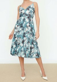 Frau in einem ärmellosen, blauen, blumenmuster-midi Kleid mit Falten, kombiniert mit weißen spitzen Absatzschuhen, stehend vor einem schlichten Hintergrund.