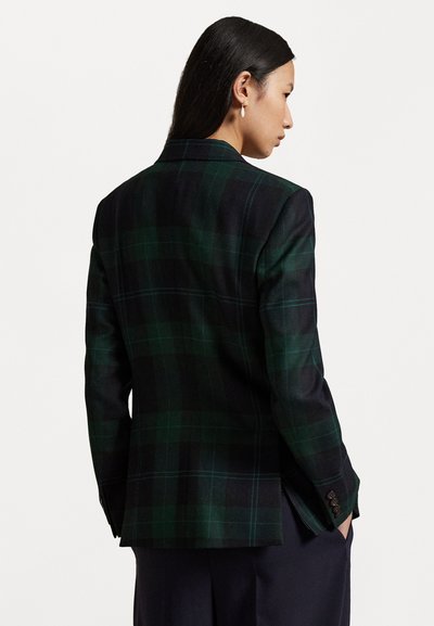 Žena s dlhými čiernymi vlasmi, oblečená v tmavozelenom a čiernom károvanom saku, stojí v profile s rukami vo vreckách námorníckych nohavíc.