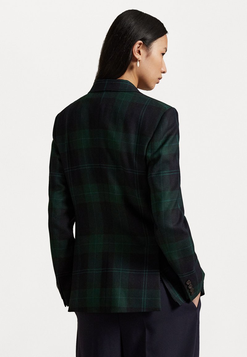 Donna con lunghi capelli neri che indossa un blazer a quadri verde scuro e nero, in posa di profilo con le mani nelle tasche dei pantaloni blu navy.