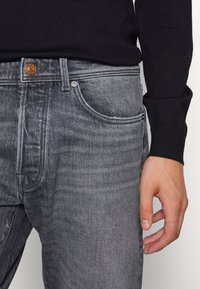 Grå denimjeans med en klassisk femficksdesign, utrustade med en kopparknapp och mjuka slitningar fram.
