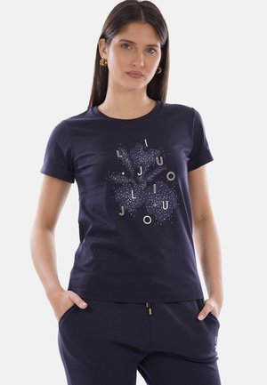 Femme aux cheveux longs et foncés portant des boucles d'oreilles créoles dorées et un t-shirt bleu marine avec un motif floral abstrait et des lettres à l'avant.