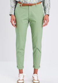 Chinos vert clair avec une coupe ajustée, featuring des poignets retroussés et une ceinture tressée beige. Associés à des baskets blanches et une chemise à manches longues à motifs.