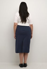 Une jupe midi bleu marine avec une fente à l'arrière, fabriquée en tissu doux, associée à un top blanc à manches courtes et des chaussures noires épaisses.