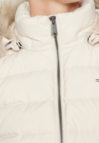 Pehmustettu kermajakku, jossa on korkea kaulus, hopeinen vetoketju ja irrotettava tekoturkisviiste hupussa. Ominaisuuksiin kuuluu vaakasuora ompelu ja hillitty logokoriste.