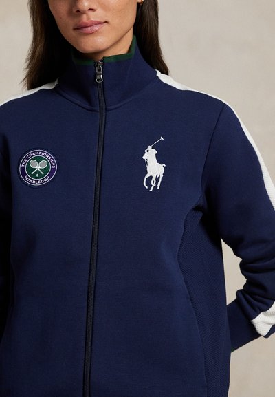 Mörkblå zip-up-jacka med vita sidostrippar, en Wimbledon-märke och en vit polo-spelare-logo. Mjukt tyg med texturerade detaljer.