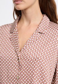 Koszula z kołnierzykiem w jasnobeżowym materiale z geometrycznym wzorem w brązowych i różowych tonach. Posiada listwę na guziki i gładką teksturę.