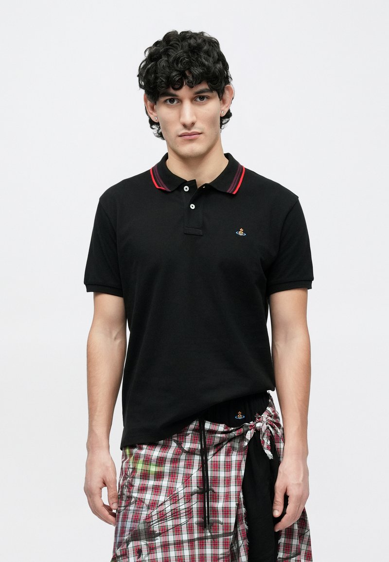 Ung mand med krøllet sort hår iført en sort poloshirt med røde striber på kraven og et ternet omslagsnederdel bundet i taljen.