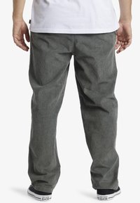 Hombre con pantalones de pana grises holgados y zapatillas negras Converse, visto desde atrás, de pie contra un fondo blanco.