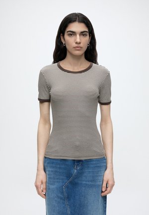 Jeune personne aux longs cheveux foncés portant un t-shirt rayé marron et blanc à manches courtes et une jupe en jean bleue, debout devant un fond neutre.