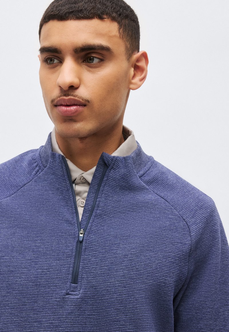 Jeune homme aux cheveux courts et bouclés portant un pull texturé bleu zippé par-dessus une chemise beige à col, regardant sur le côté.