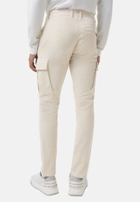 Blick von hinten auf eine Person, die beige Cargo-Hosen mit Seitentaschen und weiße Sneaker mit Branding an den Fersen trägt.