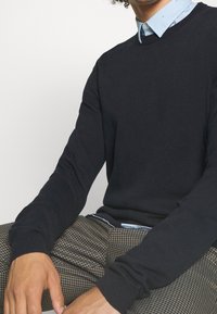 Maglione navy con col giro e polsini a coste, abbinato a una camicia colletto azzurra e pantaloni a scacchi. Tessuto morbido.