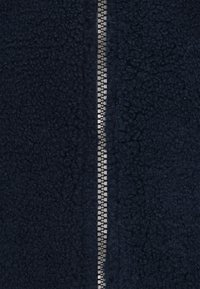 Marineblå fleecejakke med tekstureret stof, der har en central metallisk lynlås og en blød, fyldig overflade.