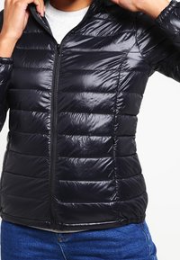 Sort dunjakke med lodret quiltet design, blank tekstur og lynlås foran. Har hætte og tilpasset talje.