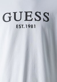 Bílá bavlněná tričko s tučným černým textem "GUESS" a "EST. 1981" umístěným uprostřed na přední straně, hladká textura a design s kulatým výstřihem.