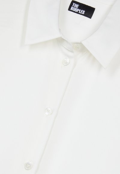 Witte button-up shirt met een gespreide kraag, parelknopen en een zwarte label met "THE KOOPLES" aan de hals. Glad textuur van de stof.