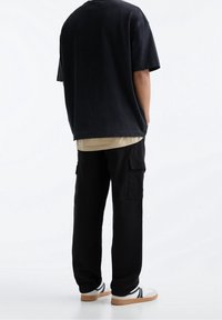 Sort oversized t-shirt med korte ærmer lagd over en lys beige bluse med lange ærmer, parret med sorte cargo bukser og hvide sneakers.