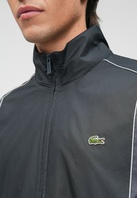 Μαύρο αθλητικό μπουφάν με φερμουάρ και ψηλό γιακά, με λευκές ρίγες και πράσινο λογότυπο κροκοδείλου στο αριστερό στήθος. Λείος υφασμάτινος σχεδιασμός.