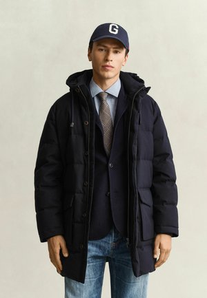 Junger Mann trägt eine dunkle Daunenjacke über einem Anzug, hellblauem Hemd, gemusterter Krawatte, blauen Jeans und einer navyblauen Mütze mit weißem Buchstaben G.