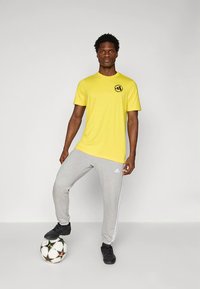 Gul kortærmet sportst-shirt med sort logo, kombineret med grå joggingbukser med hvide sidestriber og sorte sportssko, stående på en fodbold.