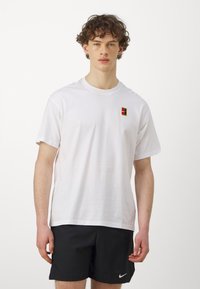 T-shirt bianca in cotone a maniche corte con un piccolo logo ricamato rosso, giallo e nero sul lato sinistro del petto. Design semplice.