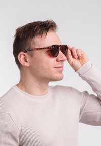 Kulaté sluneční brýle ve skořápce s tmavými skly a tenkými nohami. Nosené osobou v světlém oblečení a s krátkými, strukturovanými vlasy.