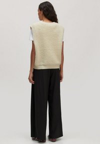 Frau mit langen dunklen Haaren trägt einen ärmellosen cremefarbenen strukturierten Sweater-Weste, ein weißes Kurzarmhemd, weite schwarze Hosen und genietetes Schuhwerk und steht mit dem Rücken zur Kamera.