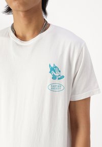 Maglietta bianca in cotone con una grafica blu di un personaggio stilizzato e il testo "KAOTIKO SOCIETY" in un design circolare sul petto.