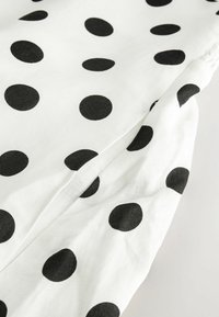 Tissu blanc à gros pois noirs, avec un détail froncé sur un bord, sur un fond clair uni.