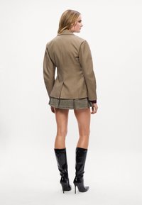 Blazer castanho com corte ajustado, combinado com uma saia mini xadrez verde e botas altas de salto stiletto pretas. Texturas de tecido suaves.