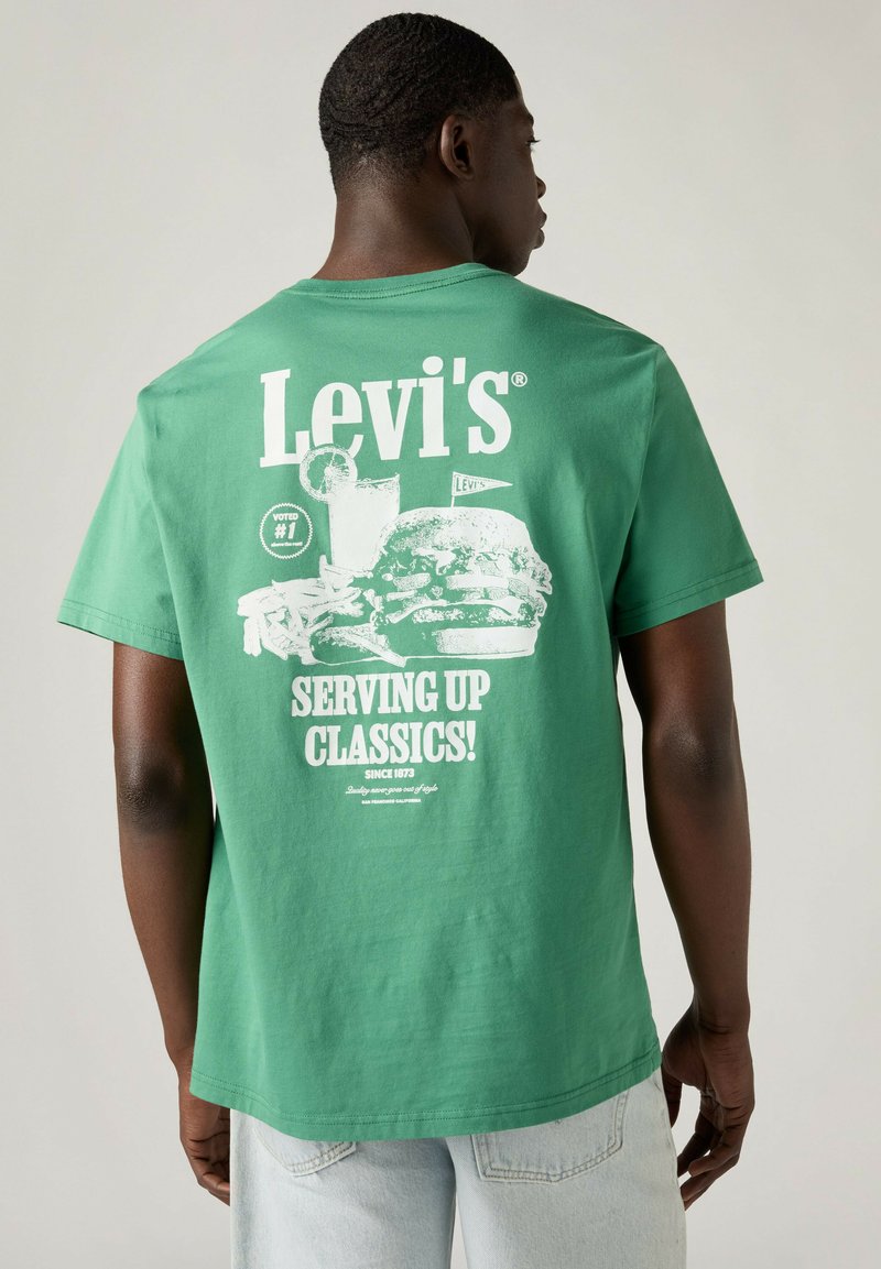 Mann trägt grünes Levi's T-Shirt mit weißer Grafik von Burger, Pommes und Getränk sowie dem Text „Serving Up Classics! Since 1873“ auf dem Rücken.
