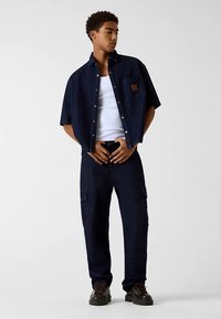 Jeanshemd mit einer Brusttasche, kombiniert mit dunkelblauen Cargohosen. Beide haben eine lockere Passform und auffällige Nahtdetails. Braune Schuhe.